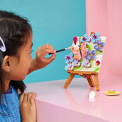 Kislány festőecsettel egy LEGO® Creator 3 az 1-ben virágos pillangó mozaikképet díszít, amely festőállványon áll. A színes építmény rózsaszín pillangót, lila virágokat és dekoratív növényi elemeket ábrázol pasztellszínű háttér előtt.