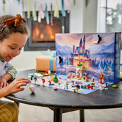 Mosolygó kislány Disney LEGO figurákkal játszik egy asztalnál, mögötte a kinyitott adventi naptár és égő kandalló.