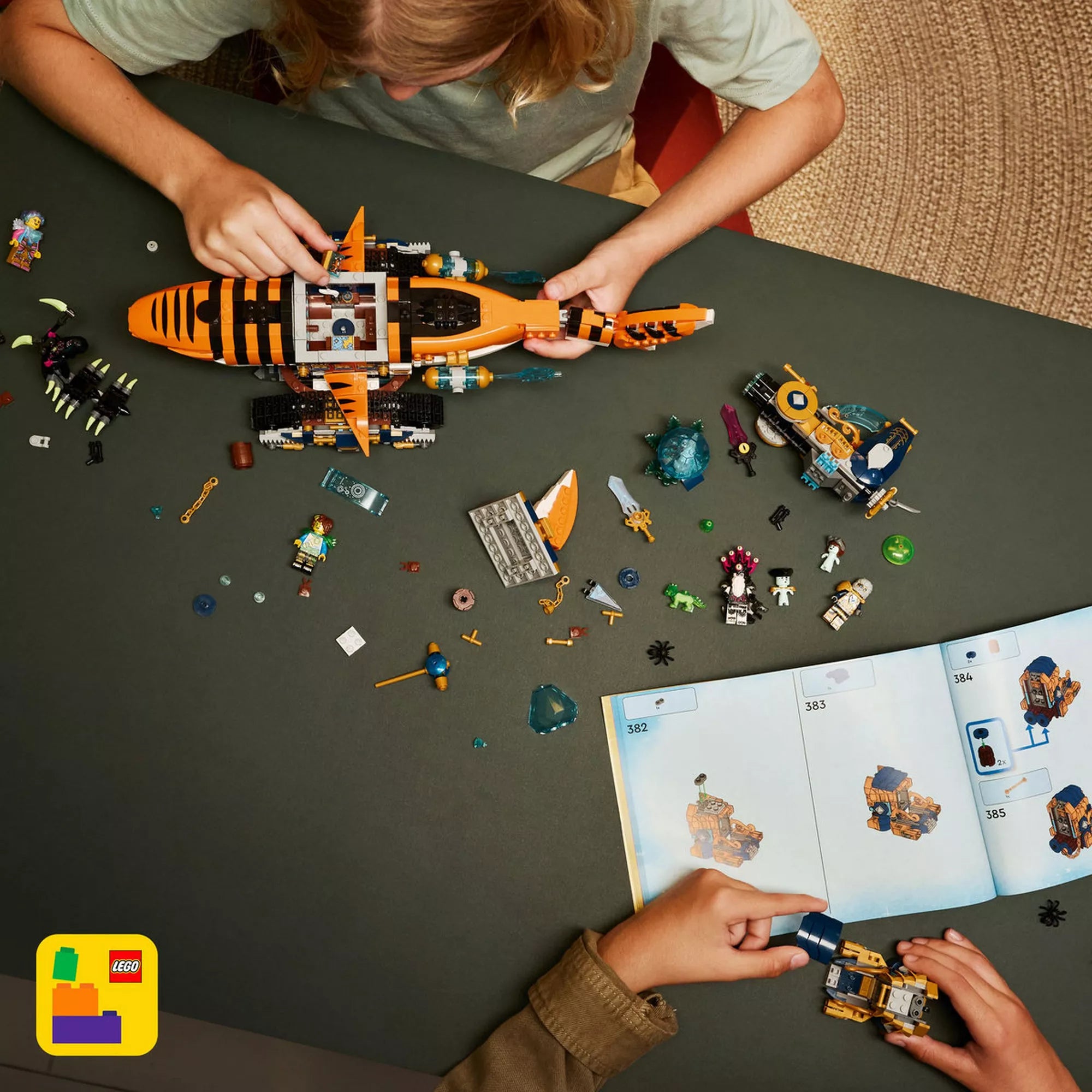 Felülnézeti kép a LEGO DREAMZzz Tigriscápatank építéséről, szétszórt elemekkel, minifigurákkal és a nyitott építési útmutatóval.