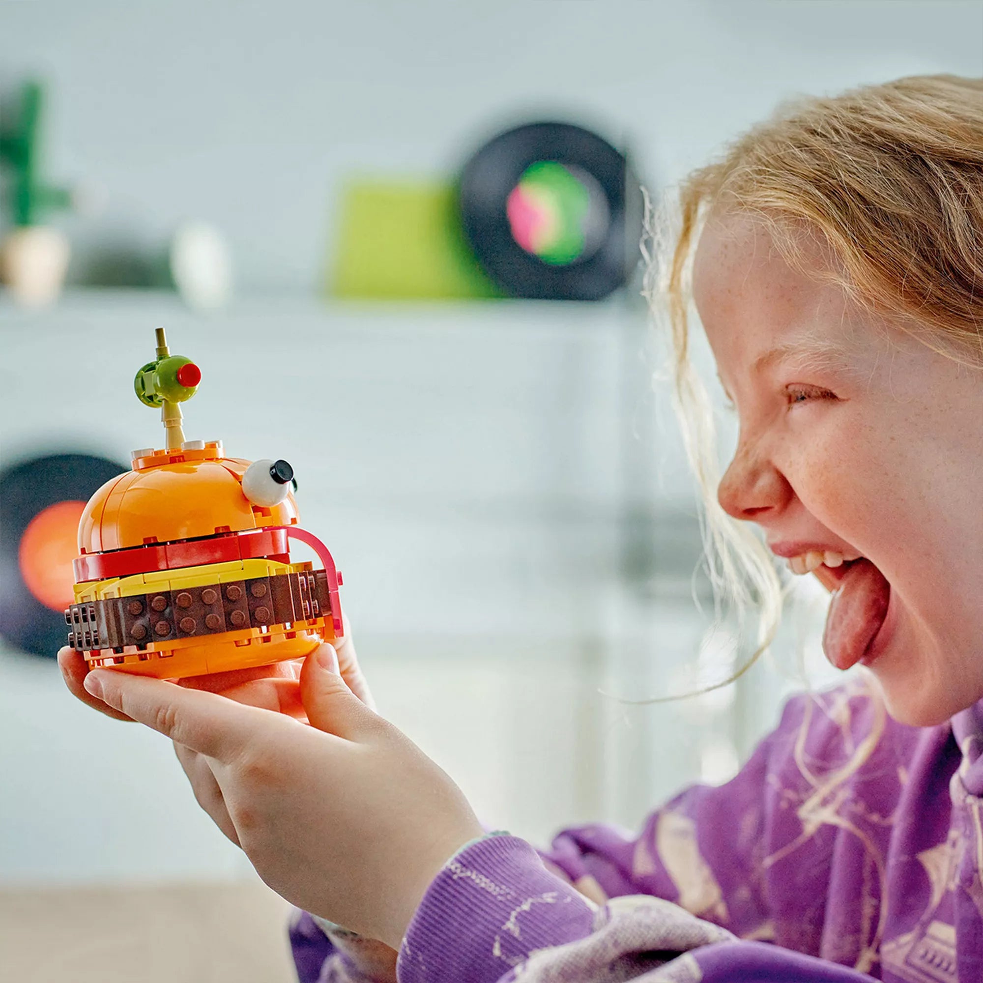 Egy mosolygó kislány kezében tartja a megépített LEGO Fortnite Durrr Burger modellt, miközben nevetve utánozza a figura kinyújtott nyelvét. A kép jól bemutatja a játék vidám hangulatát.