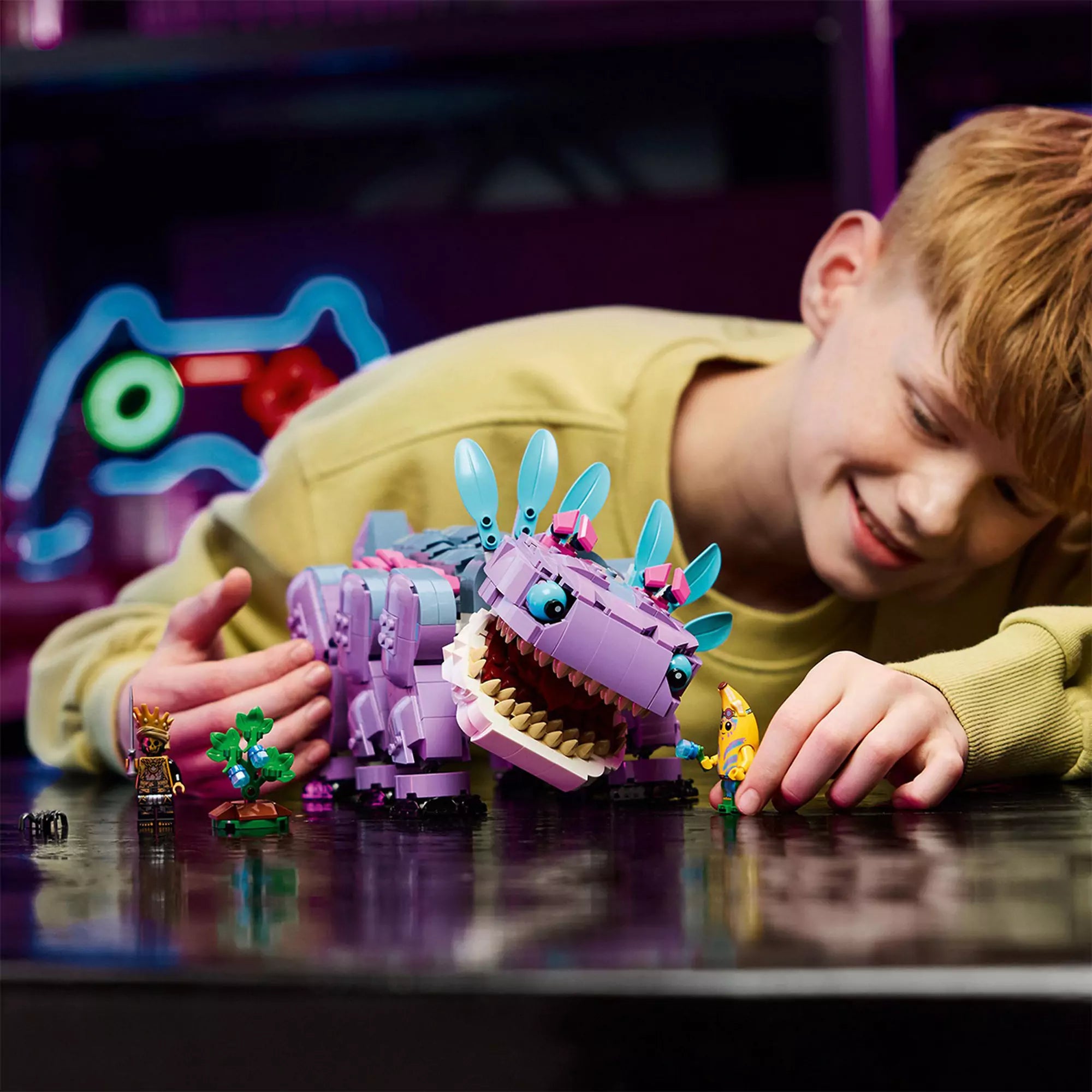 Egy mosolygó fiú játszik a LEGO Fortnite Klombo szettel: a lila szörny figura és a három minifigura együtt látható az asztalon, izgalmas játékkörnyezetben.