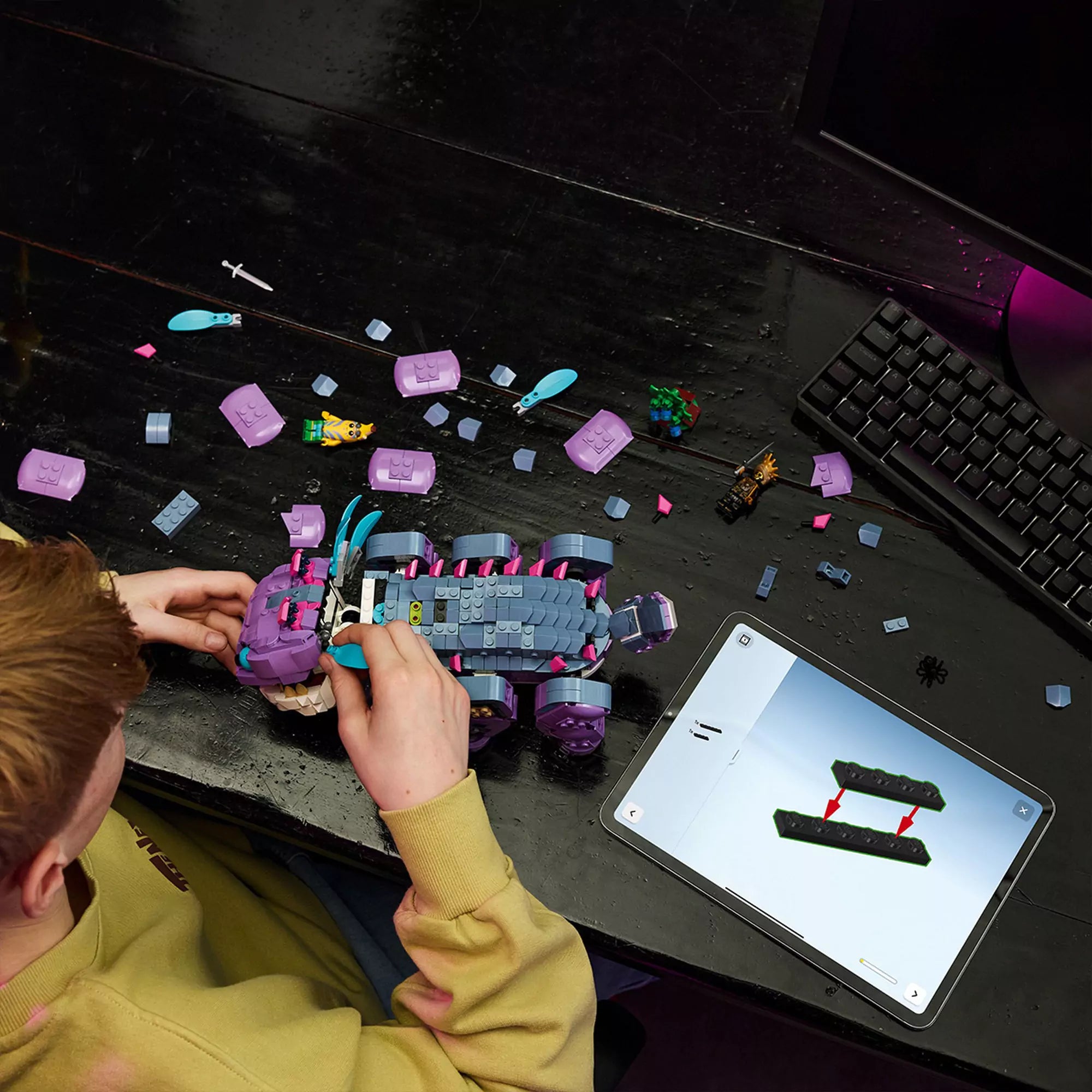 Egy gyermek építi a LEGO Fortnite Klombo lila szörnyét egy fekete asztalon, körülötte szétszórt elemek, minifigurák és egy tablet mutatja az útmutatót.