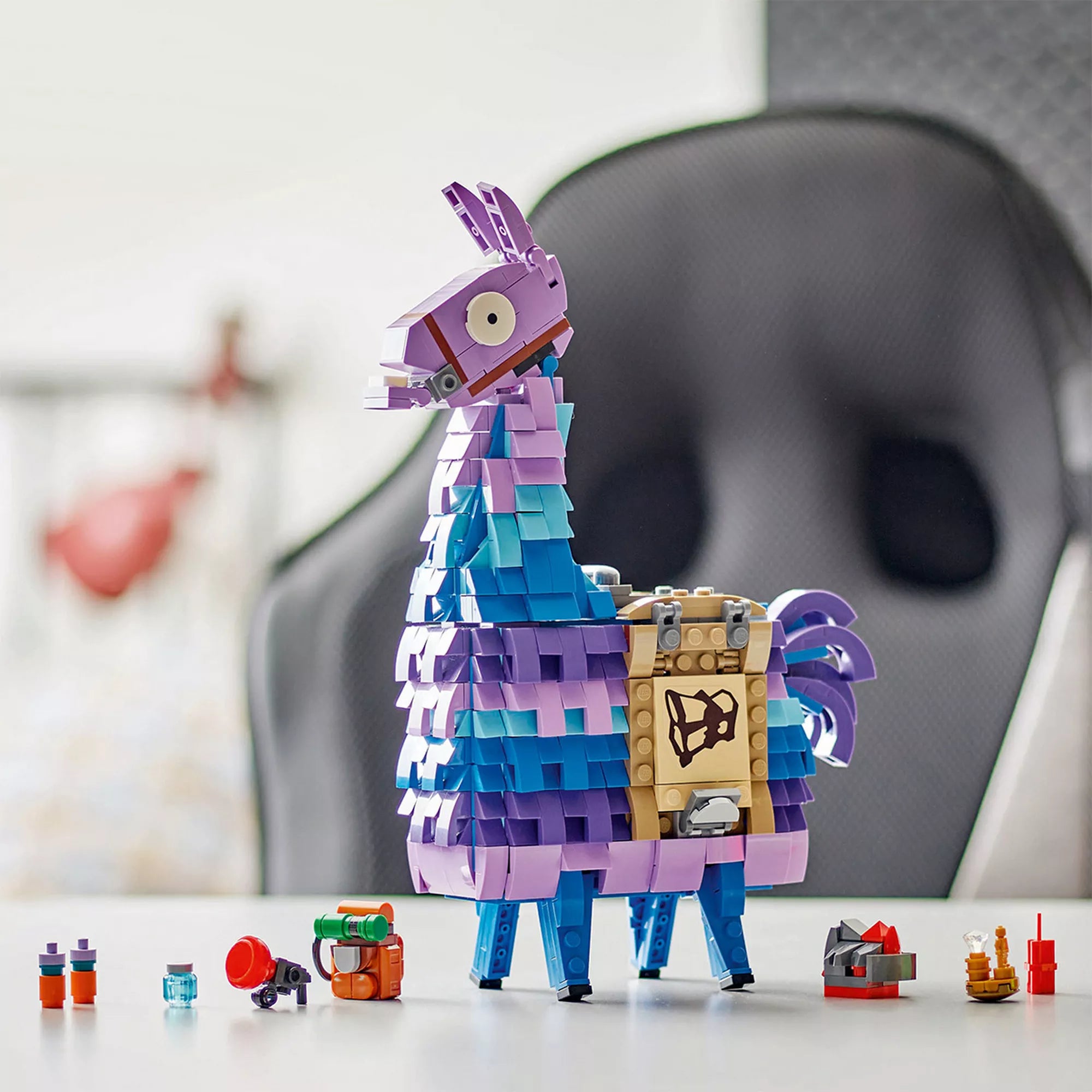 A megépített LEGO Fortnite Supply Llama figura asztalon kiállítva, a játékban ismert ikonikus színekben: lila, kék és türkiz. A láma mellett apró építhető Fortnite kiegészítők láthatók, például hátizsák, italok és dinamitrúd.