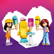 LEGO Friends Liann és Paisley minifigurák a fagylalt alakú kabalafigurával, kézben tartott fagylaltokkal és állítható asztallal, szív alakú háttér előtt.