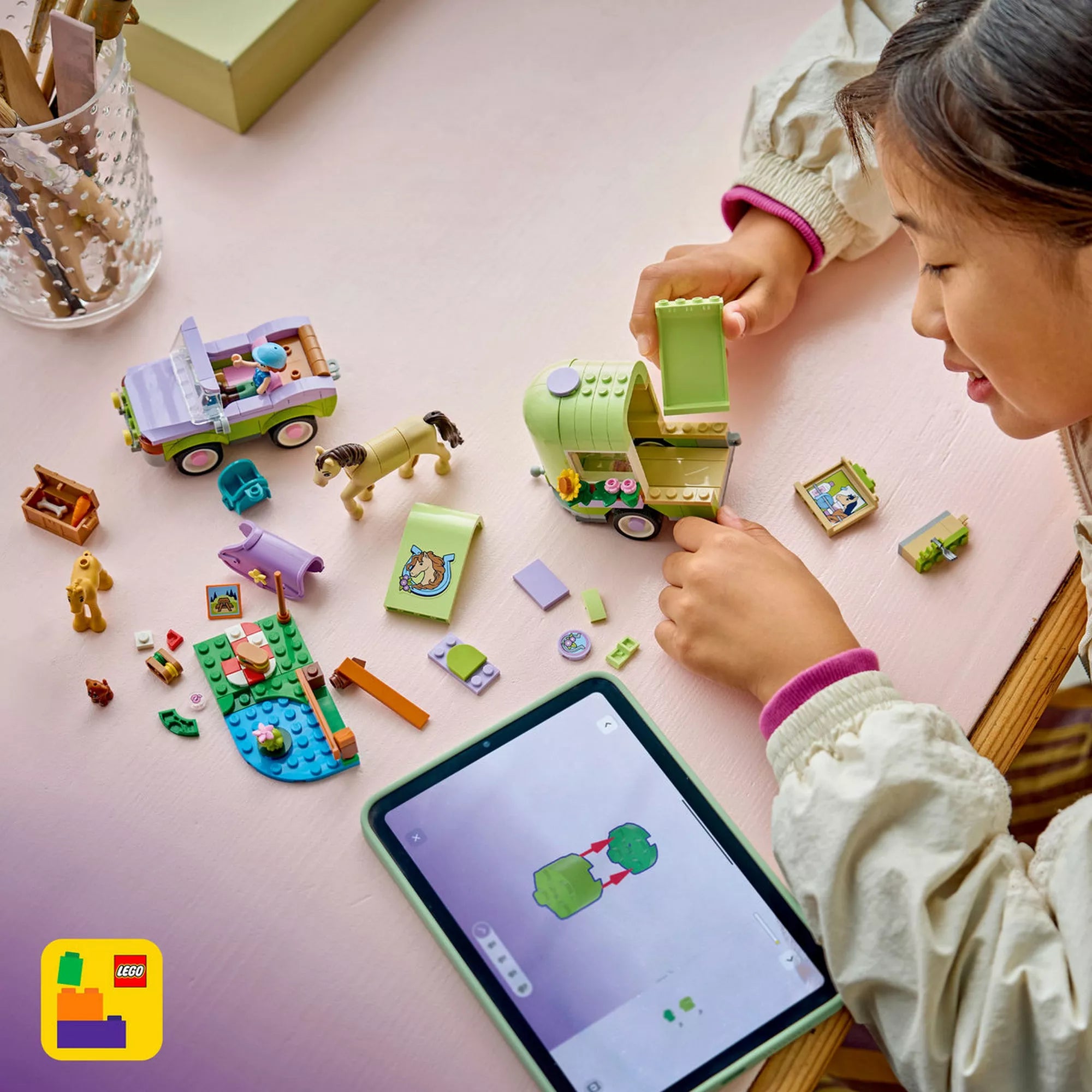 LEGO Friends Lószállító utánfutó és csikó (42695) készlet építése digitális útmutatóval, ahol a szétszedett elemek, a lószállító utánfutó és egy tablet képernyője is látható.