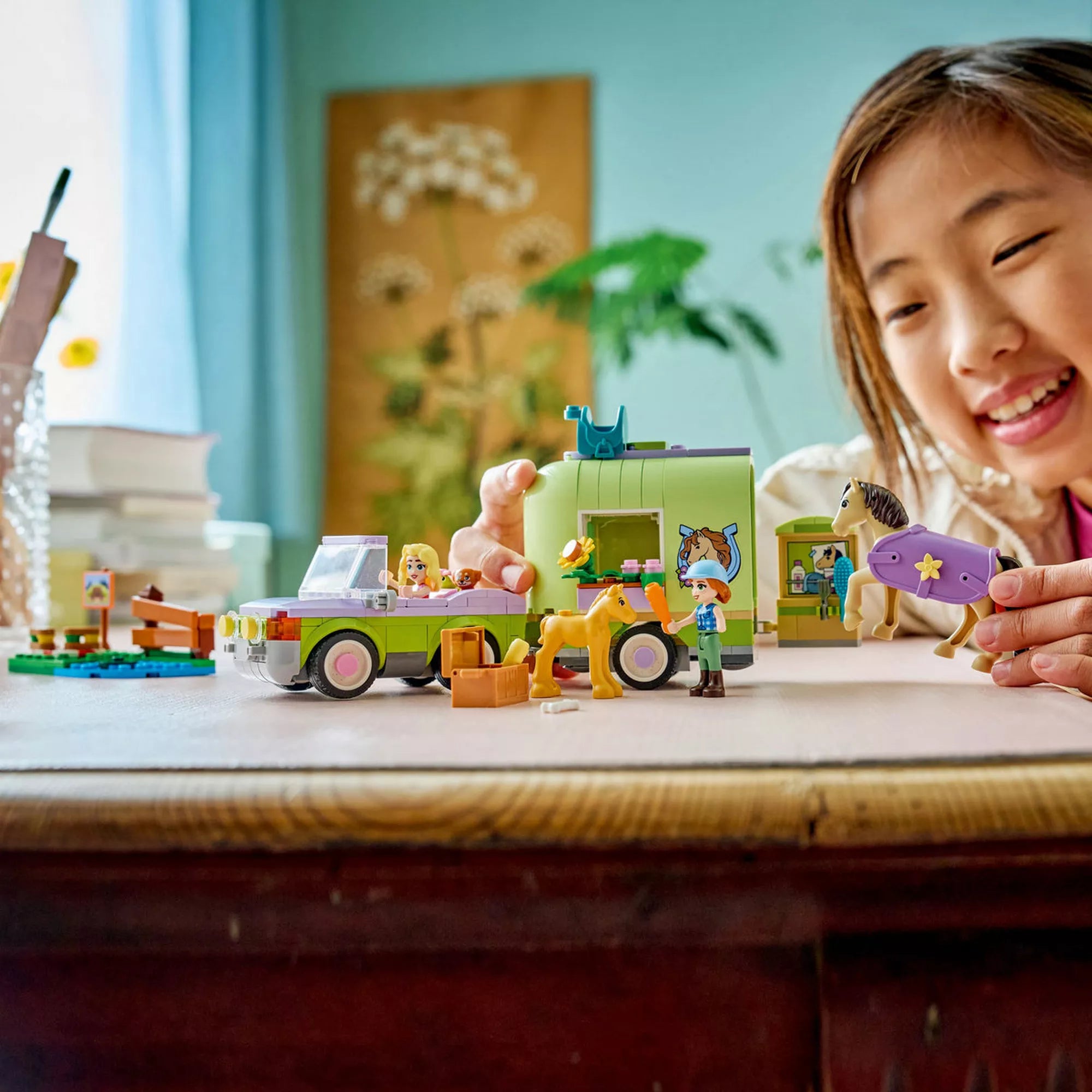 LEGO Friends Lószállító utánfutó és csikó (42695) építőjáték használat közben, ahol egy gyermek a terepjáróval, utánfutóval, lovakkal és minifigurákkal játszik egy asztalon.