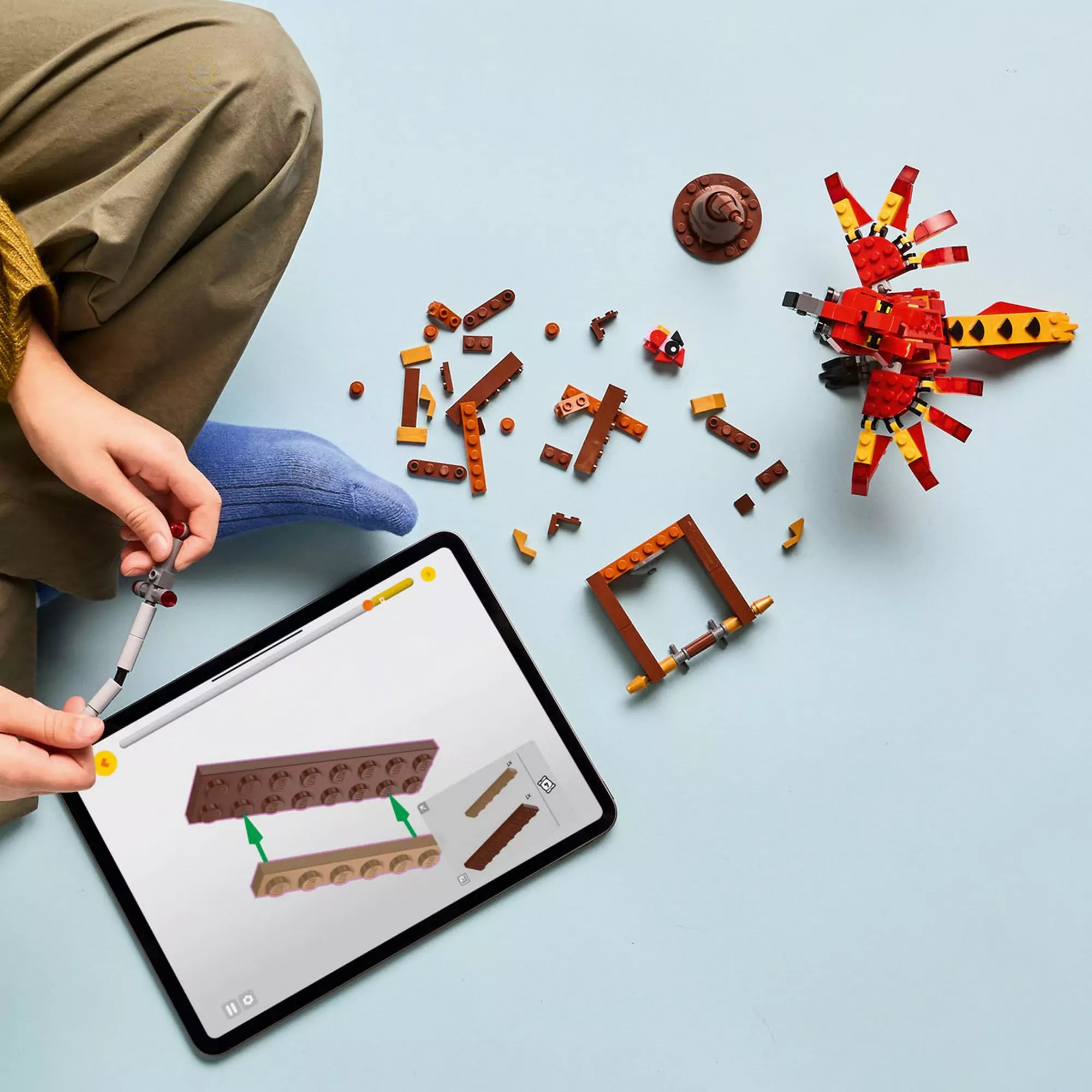 Egy gyermek építi a készletet tableten megjelenített digitális útmutató segítségével, körülötte szétszórt LEGO elemek, köztük a Fawkes figura és a barna állvány részei.