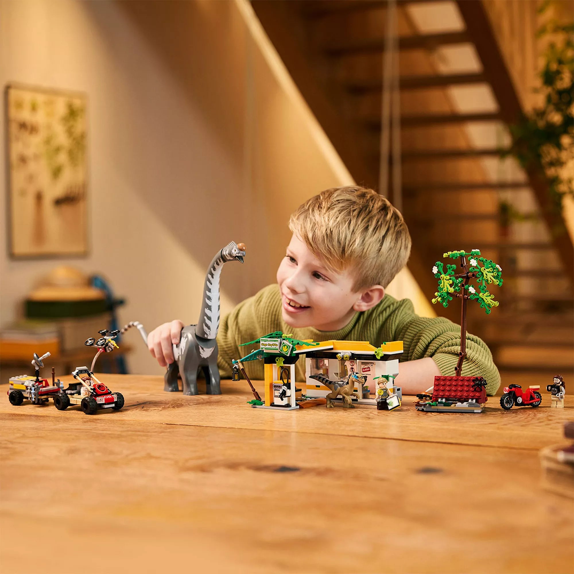 „Egy gyermek a LEGO Velociraptor és Titanosaurus üldözése készlettel játszik az asztalon: a mozgatható Titanosaurus, a tábori állomás, a járművek és a minifigurák mind jól láthatók.”