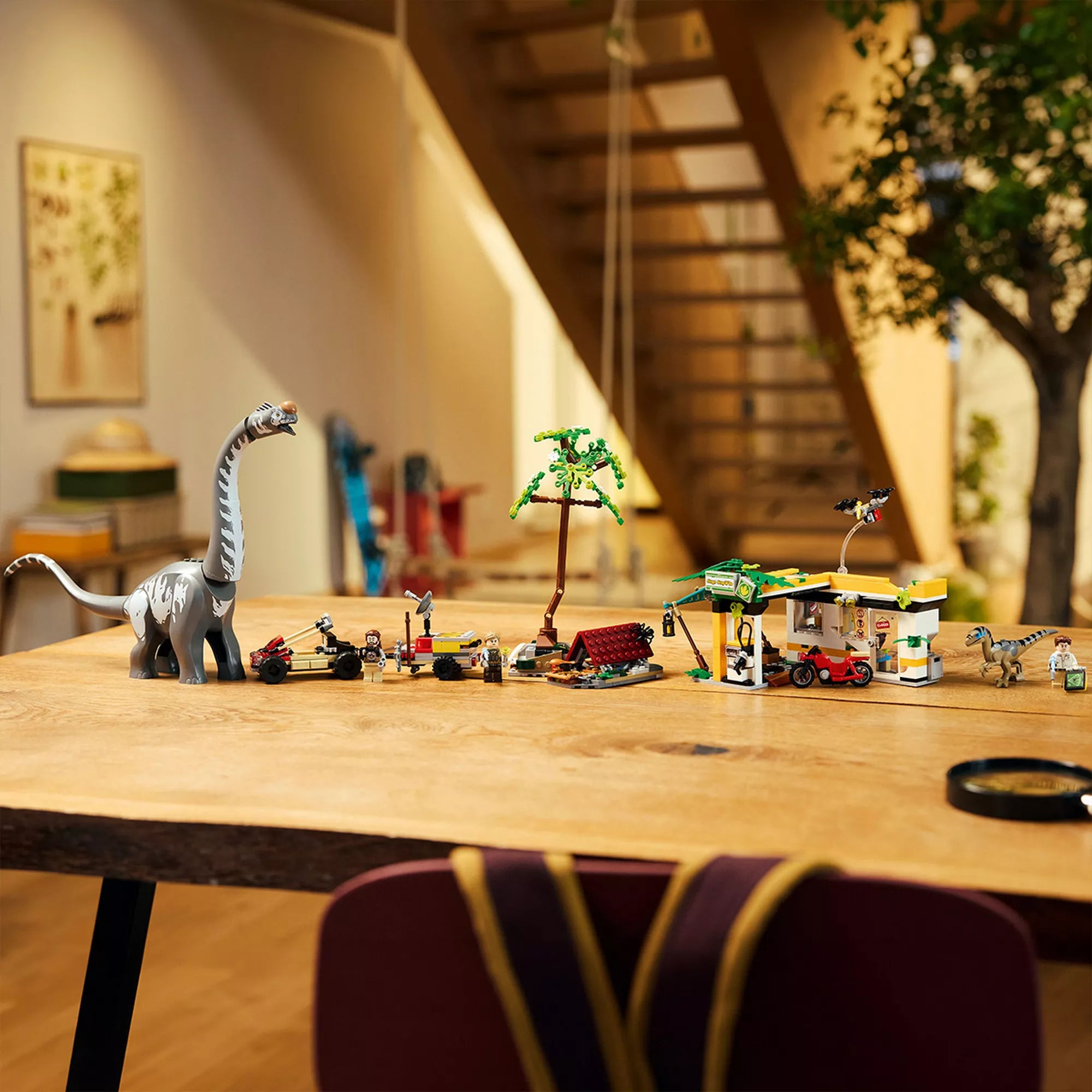 „A teljes LEGO 76973 készlet egy asztalon kiállítva: a hatalmas Titanosaurus, a Velociraptor, a terepjáró, a motor, a drón, a fa és a tábori építmény egy jelenetben.”