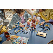 Két gyermek a LEGO NINJAGO Iker titánpáncélok 71870 készletet építi, miközben a robotfigurák, fegyverek és minifigurák az asztalon láthatók.