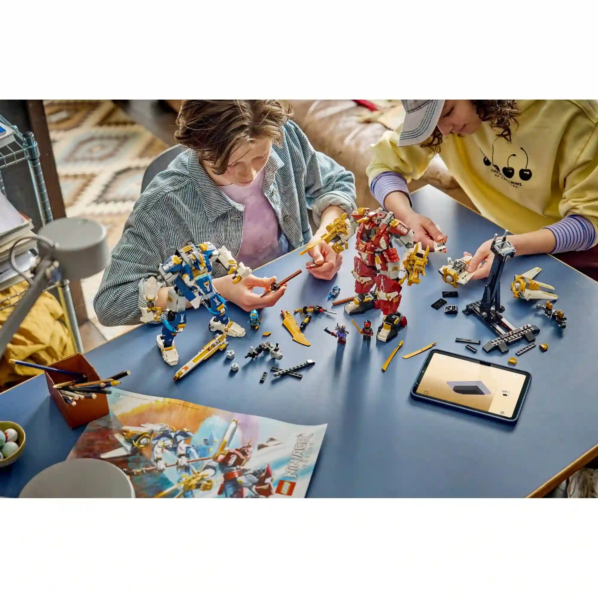 Két gyermek a LEGO NINJAGO Iker titánpáncélok 71870 készletet építi, miközben a robotfigurák, fegyverek és minifigurák az asztalon láthatók.