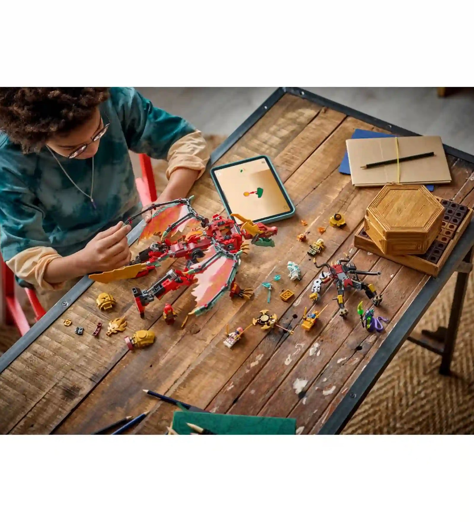 Gyermek építi és rendezi a LEGO NINJAGO Wyldfyre átalakuló sárkányrobotja 71868 készlet elemeit egy faasztalon, a háttérben LEGO Builder alkalmazással.