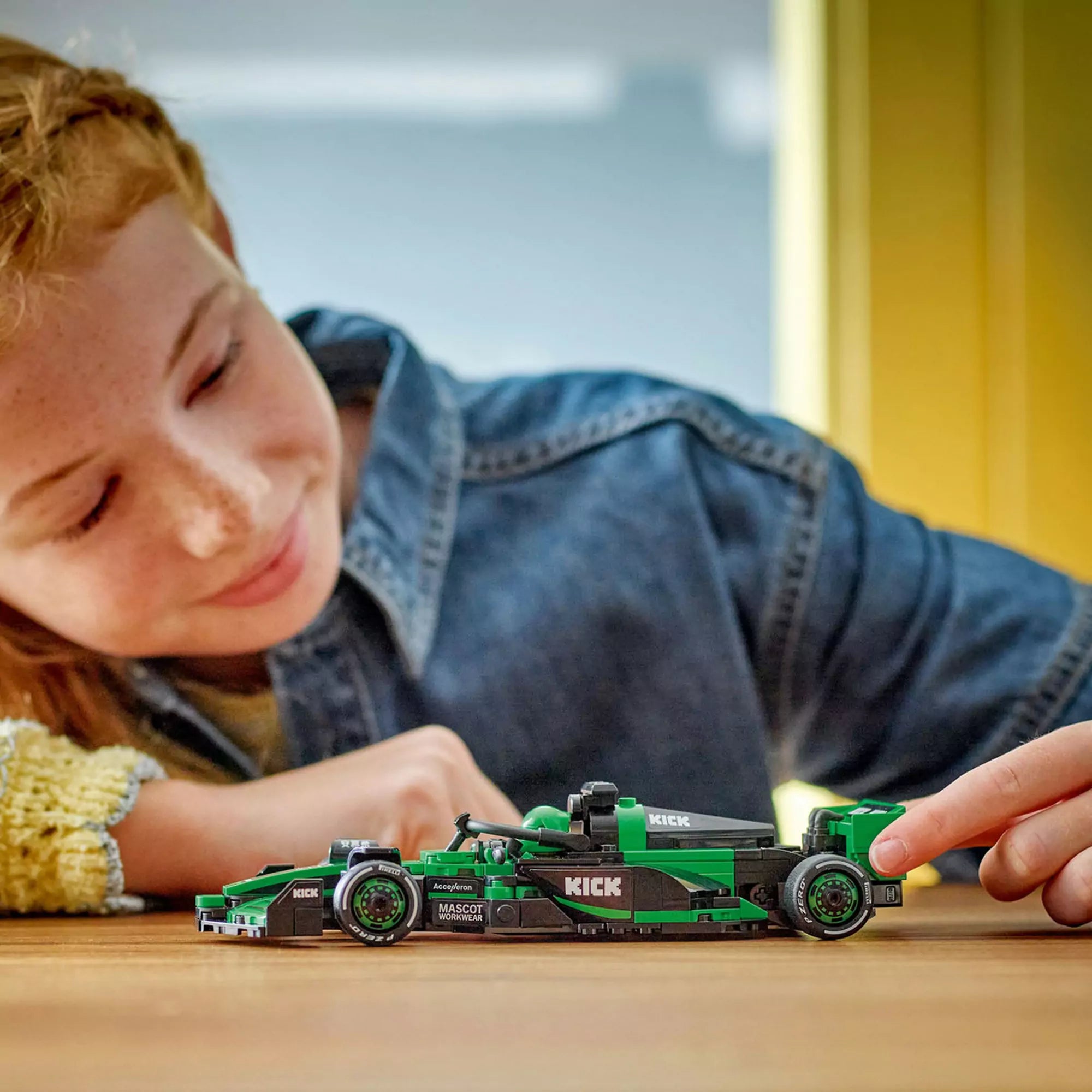  Lány az asztalnál játszik a zöld-fekete LEGO Sauber F1 autóval, oldalnézetből megörökítve.