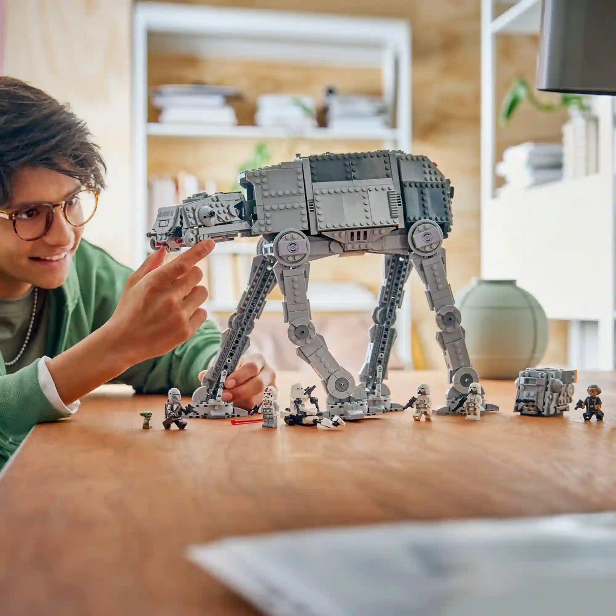 Szemüveges fiatal a szürke LEGO Star Wars AT-AT lépegető fejét mozgatja, az asztalon minifigurák és egy kisebb jármű látható.