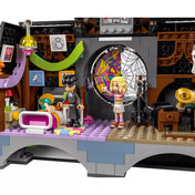 „A LEGO Wednesday és Enid kollégiumi szobájának részletes belső tere: Wednesday és Enid minifigurái a pókhálós ablak előtt, bal oldalon a színes ágy és dekorációk, jobb oldalon a cselló, gramofon és az ikonikus rózsaszín-lila színvilág.”