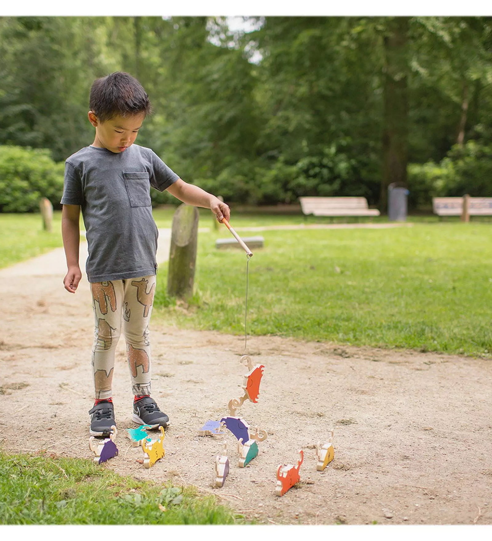 Egy kisfiú játszik kint a parkban a BS Toys Crab Fishing játékkal, színes fa rákokat emel fel egy mágneses horgászbottal a földről.