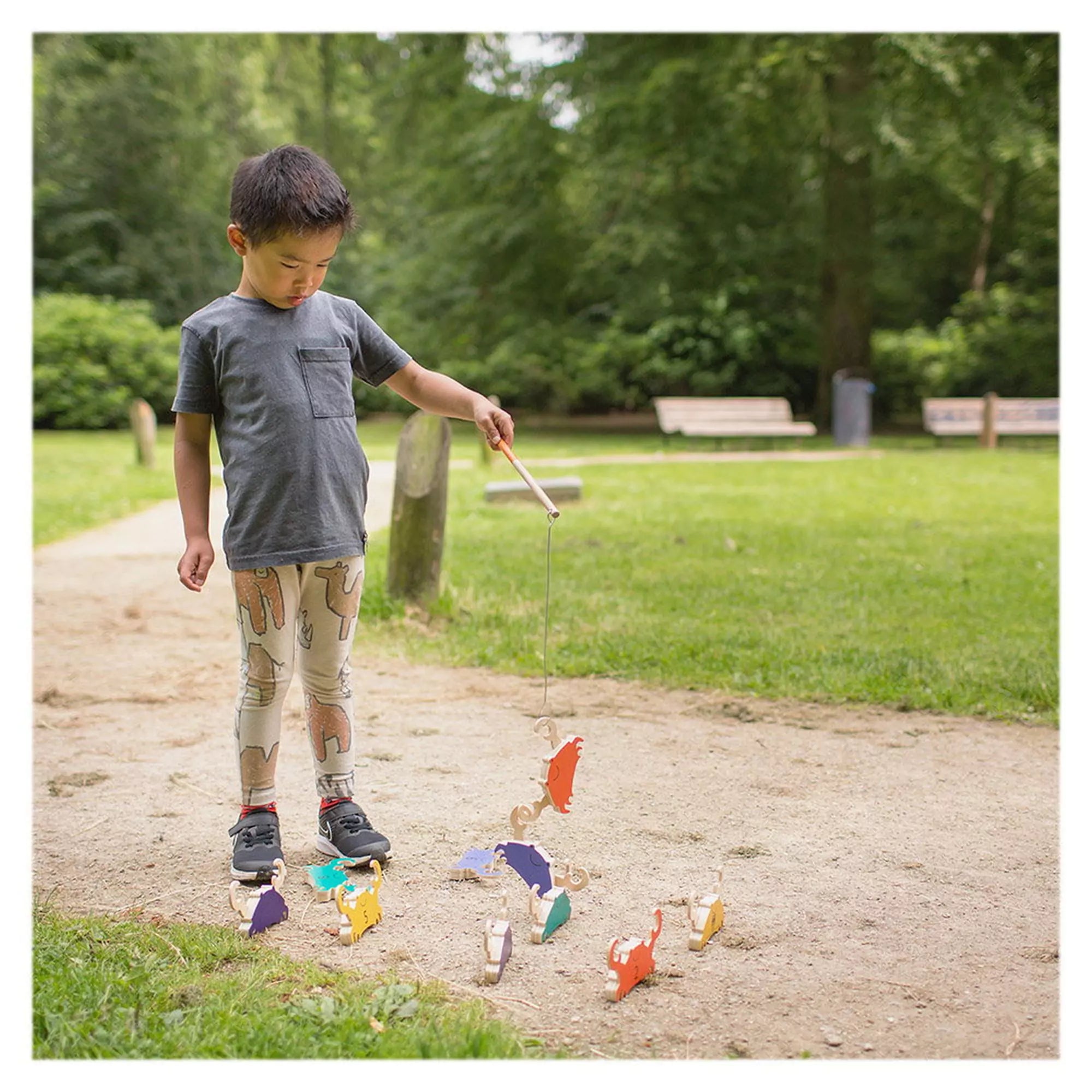 Egy kisfiú játszik kint a parkban a BS Toys Crab Fishing játékkal, színes fa rákokat emel fel egy mágneses horgászbottal a földről.