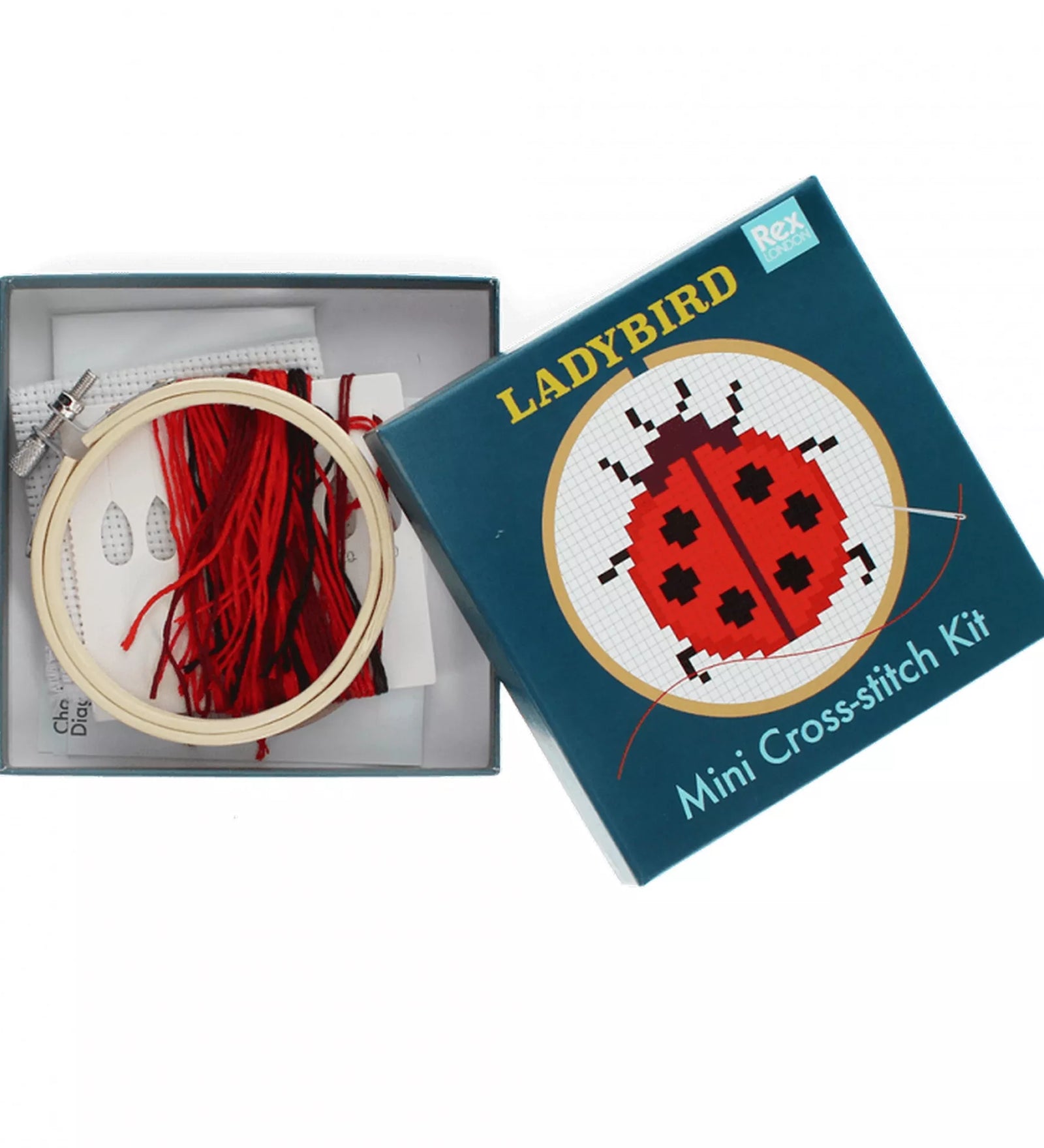 A Ladybird Mini Cross-stitch Kit nyitott doboza, benne a fa hímzőkerettel, piros pamutfonalakkal, Aida hímzőanyaggal és a mintát tartalmazó útmutatóval. A doboz fedele a katicabogár mintával a háttérben látható.