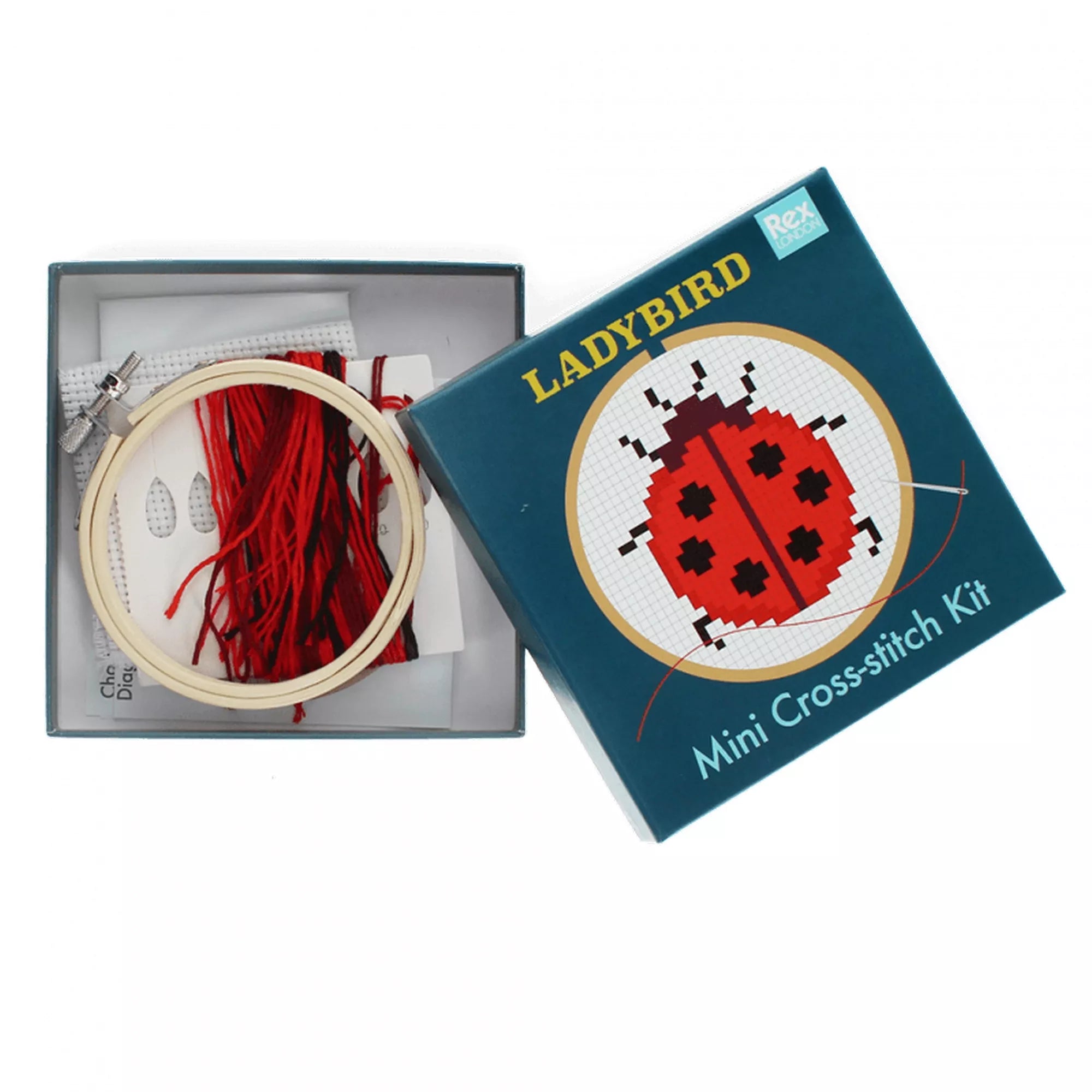 A Ladybird Mini Cross-stitch Kit nyitott doboza, benne a fa hímzőkerettel, piros pamutfonalakkal, Aida hímzőanyaggal és a mintát tartalmazó útmutatóval. A doboz fedele a katicabogár mintával a háttérben látható.