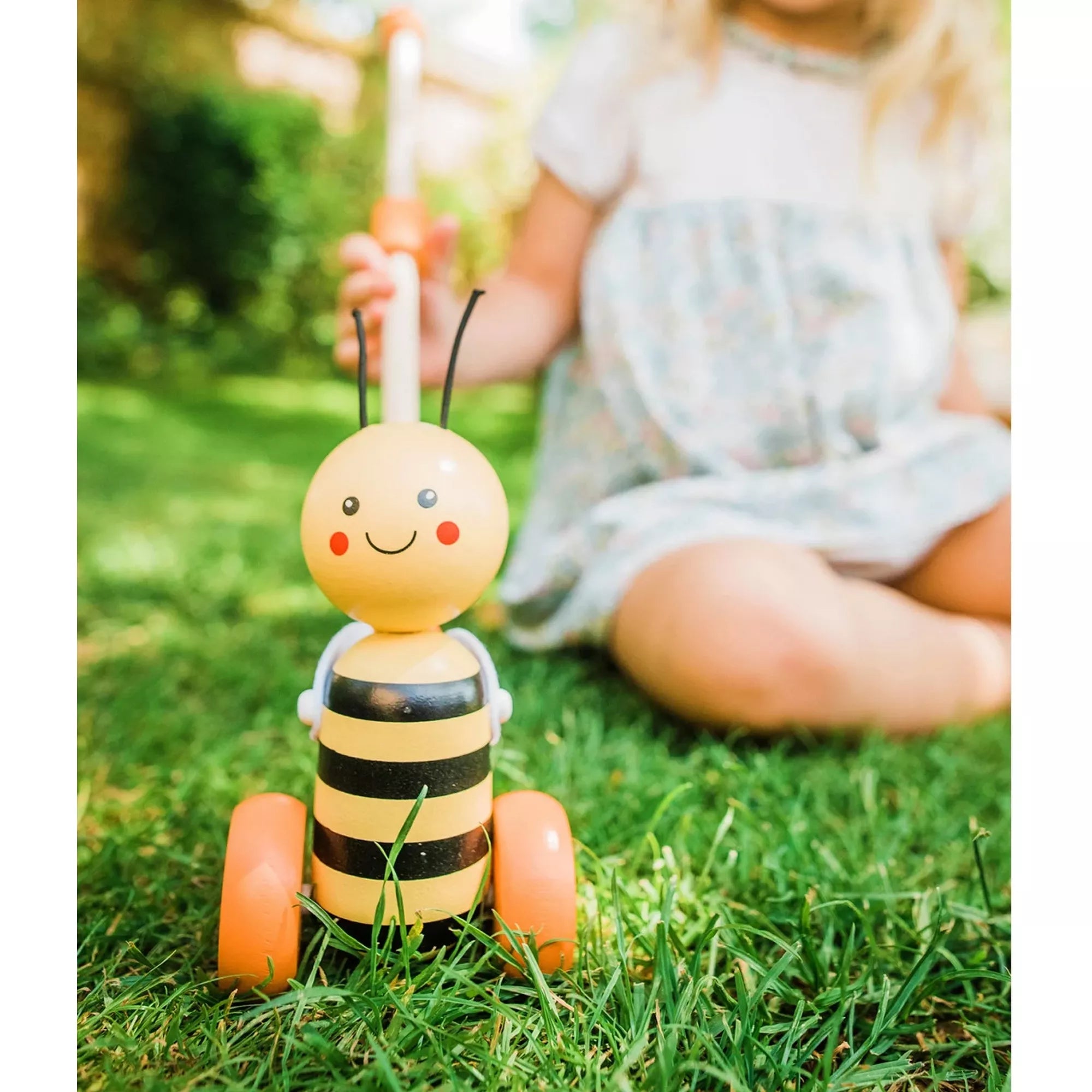A tologatható fa méhecske játék fűben, egy kisgyermek mellett. A vidám arcú, csíkos méhecske narancssárga kerekeken áll, miközben a gyermek a tolórudat fogja. A kép játékos, kültéri jelenetet ábrázol.