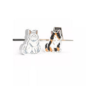 Két aranyos macskafigura a Virágvásár társasjátékból: egy fehér és egy háromszínű cica. A fafigurák részletesen illusztráltak, a háttérben a játék táblája halványan látható.